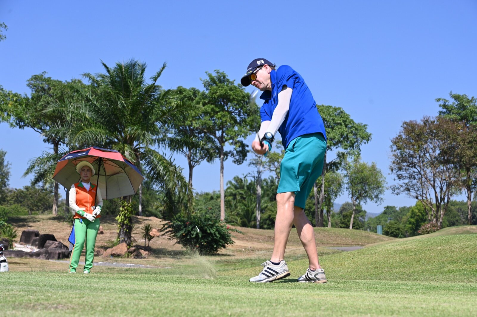 A golfer in a blue shirt hits a tee shot while a caddie in an orange bib stands under a large striped golf umbrella