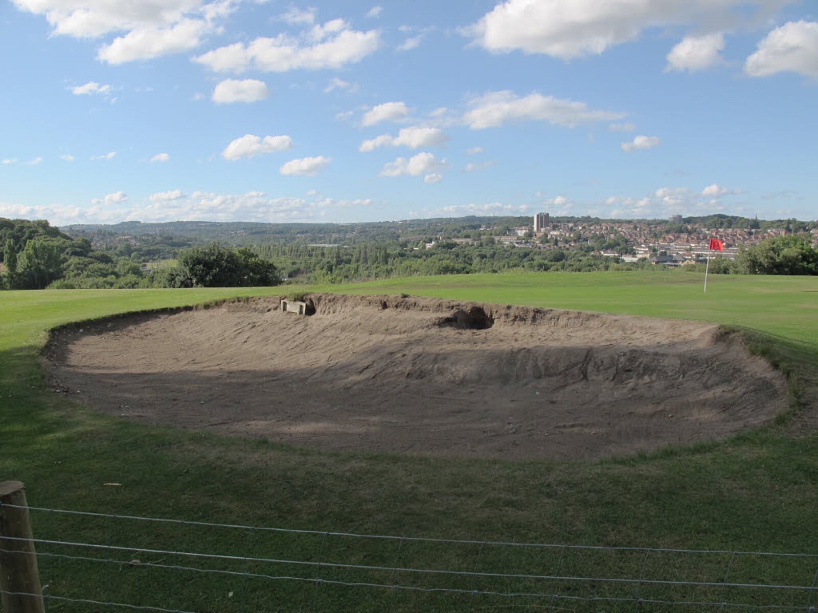A large greenside bunker with a steep sand face in front of a mown green, with a red flagstick beyond