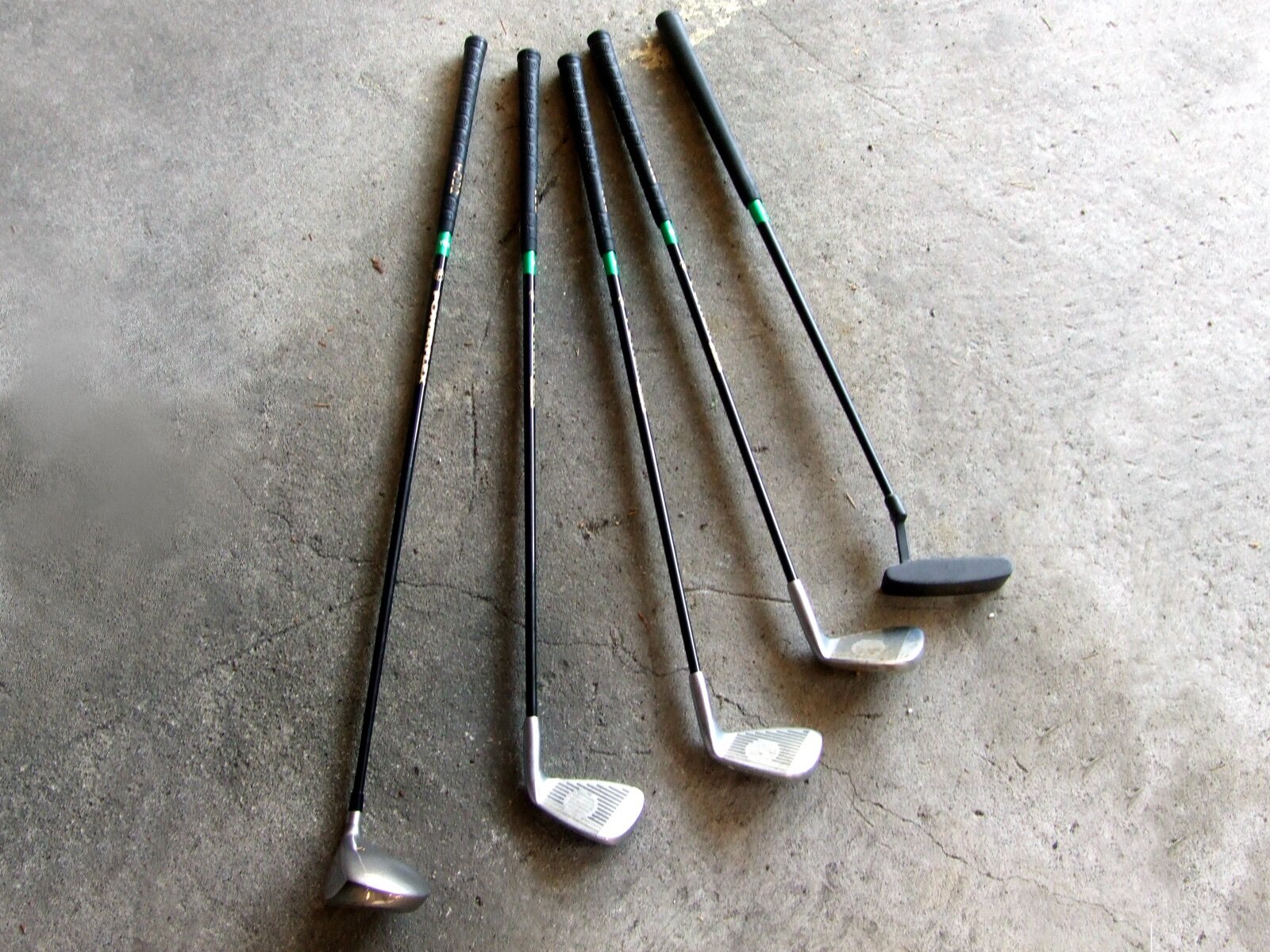 A small set of junior golf clubs laid out on concrete: woods, irons and a putter