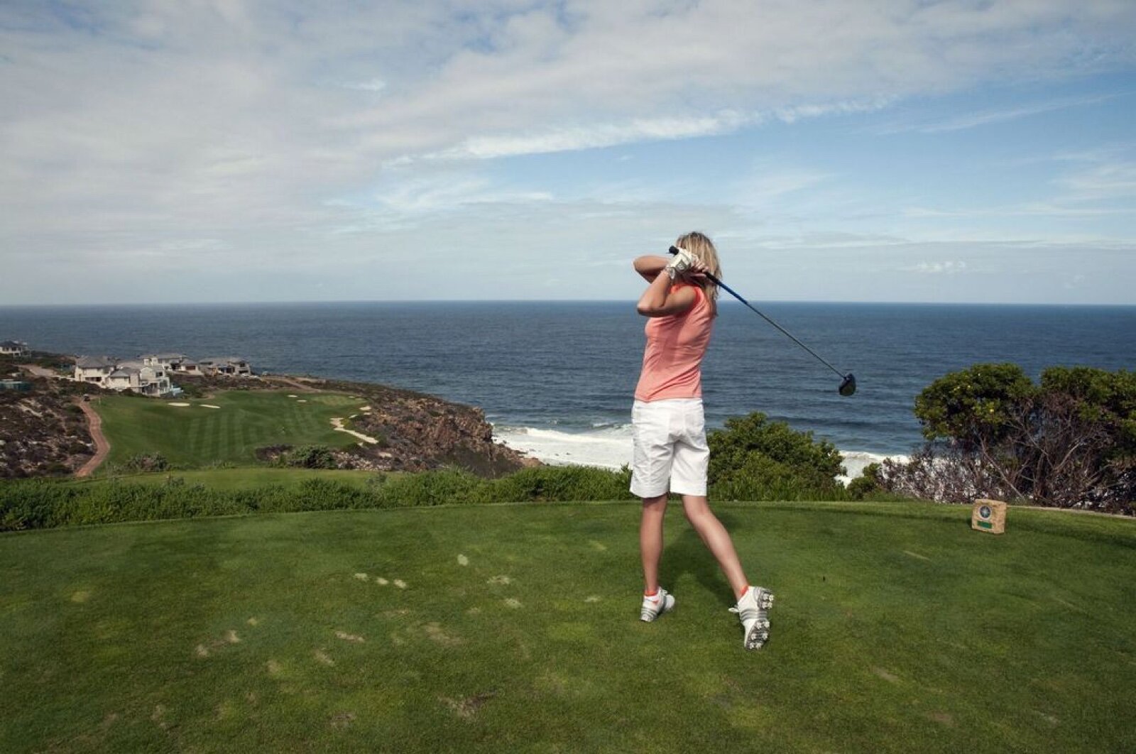 A woman golfer in a pink top finishing her swing with a driver on a clifftop tee above the sea