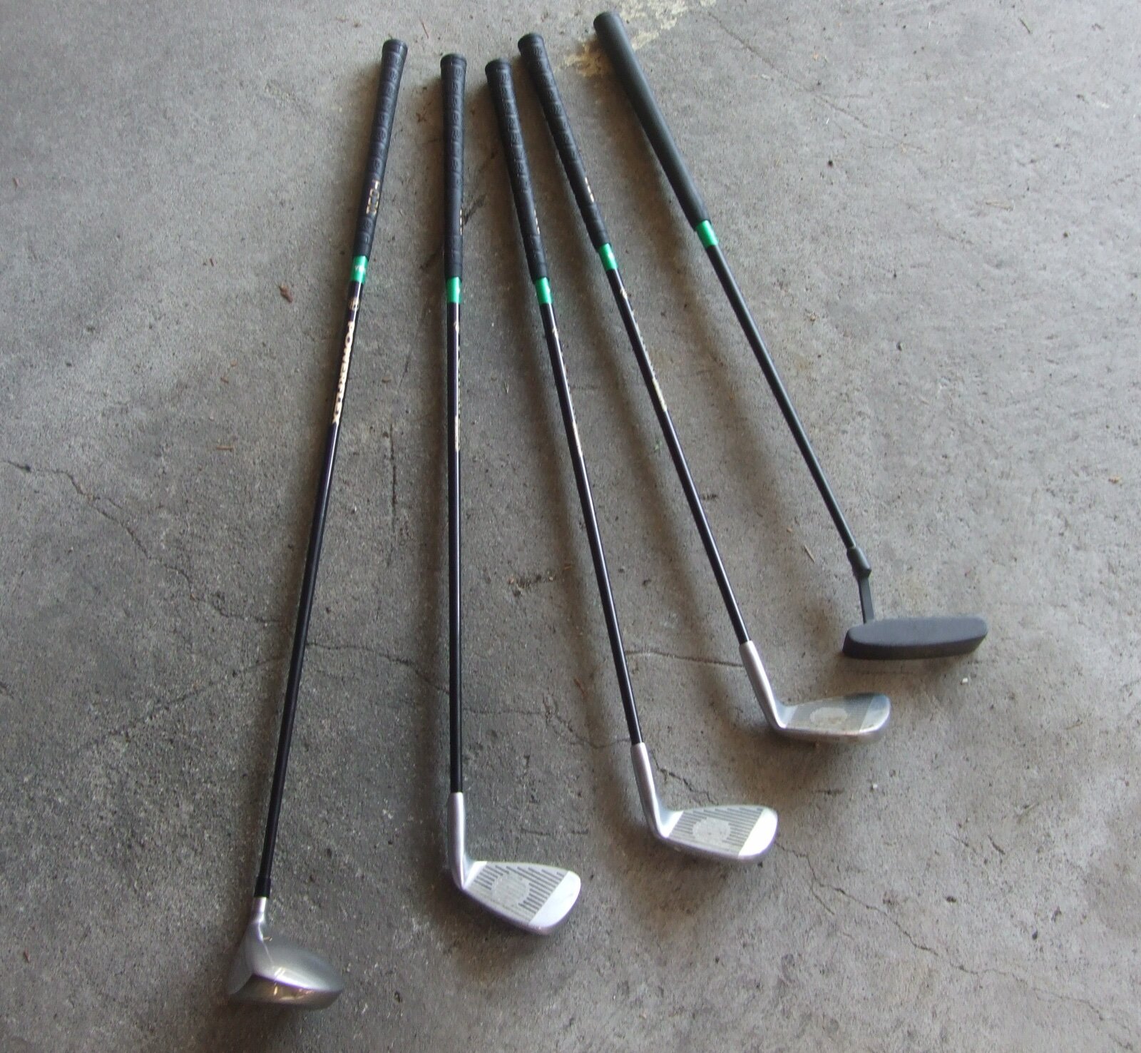 A junior set of left-handed golf clubs, four irons and a putter, laid out on a concrete floor