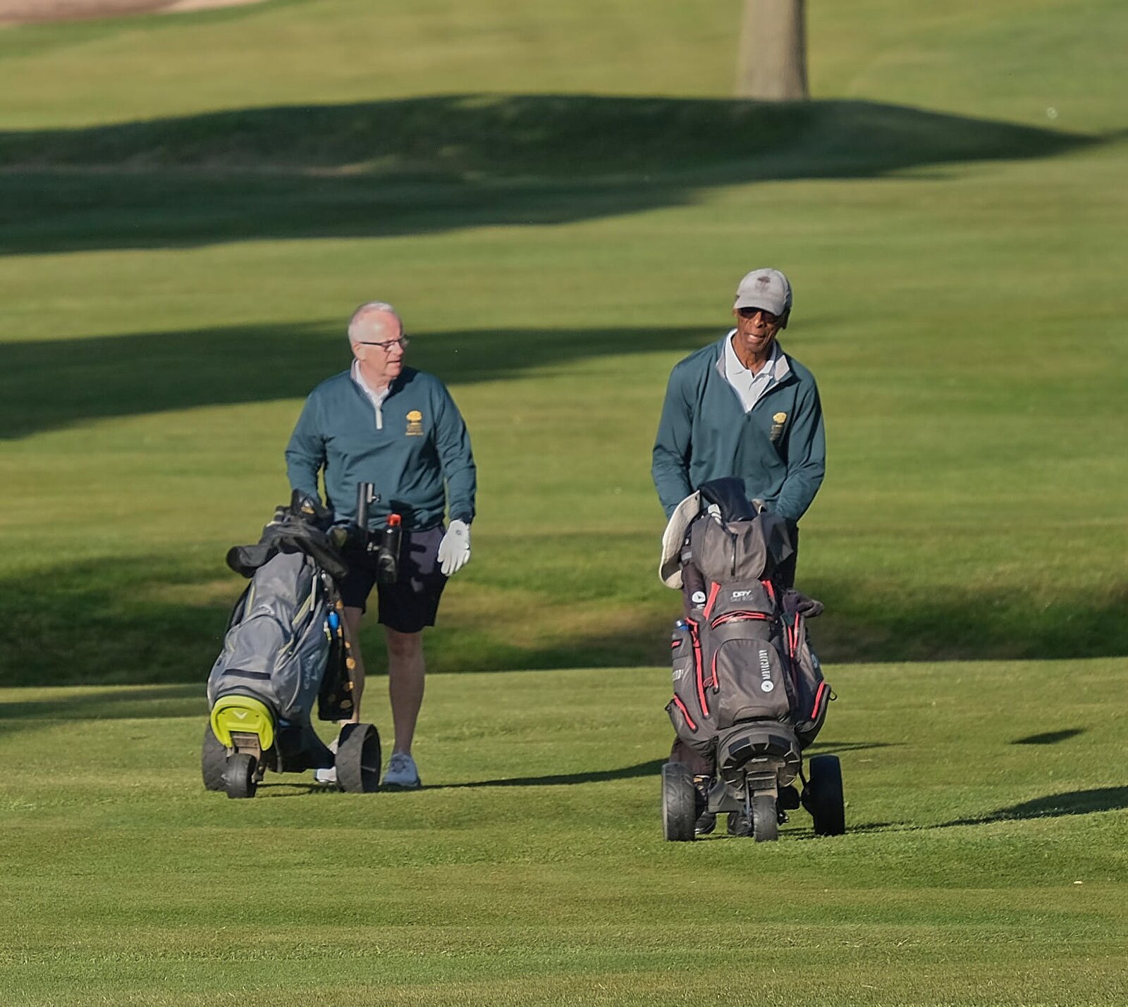 Two golfers walking across a fairway, each with a golf bag on a three-wheeled trolley