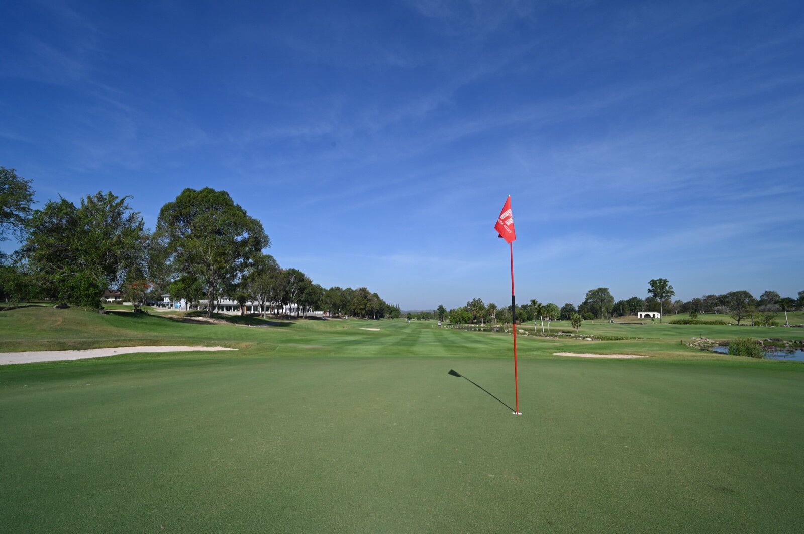 A golf green with a red flag in the hole and the fairway stretching away under a blue sky