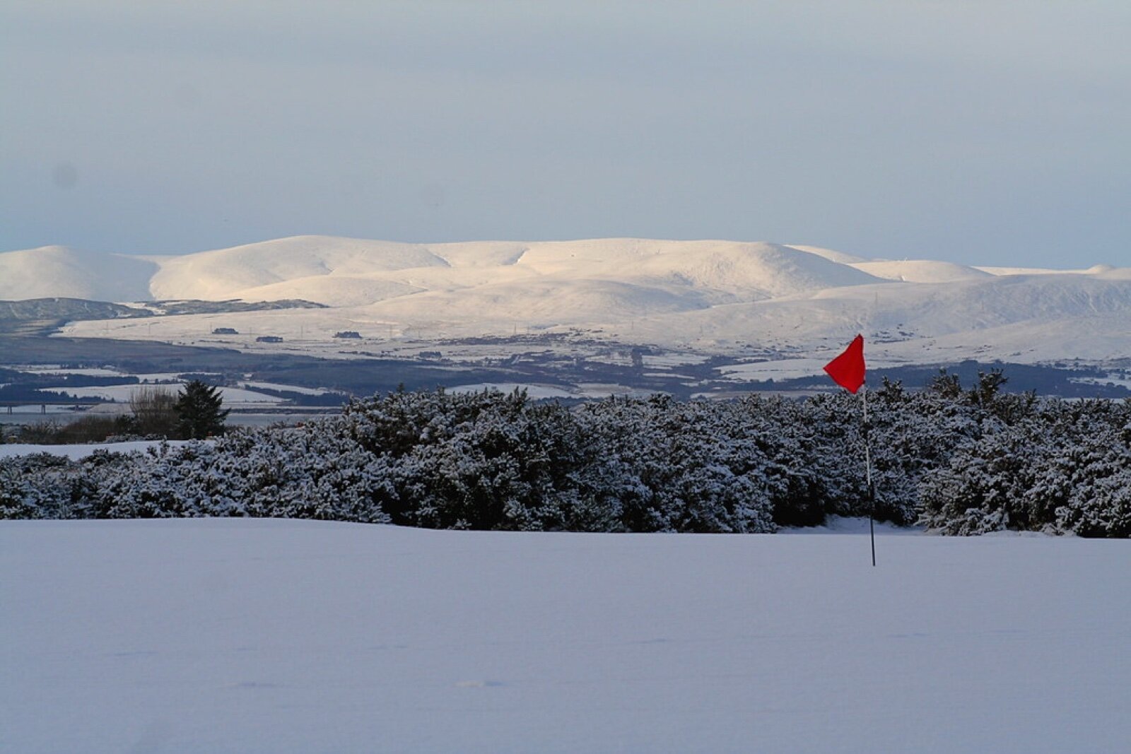 A snow-covered golf green with a red flagstick, hills behind, at Royal Dornoch Golf Club in winter