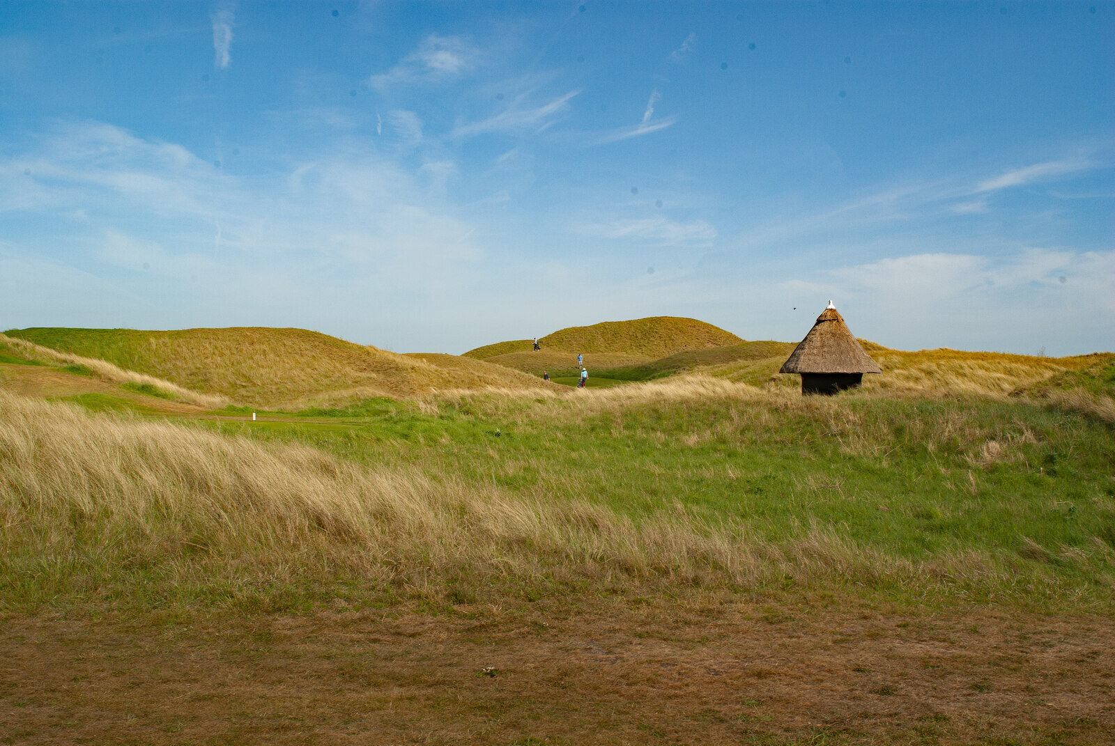 Dunes and rough on the Royal St George's golf links at Sandwich, with a thatched shelter on the right and golfers in the distance